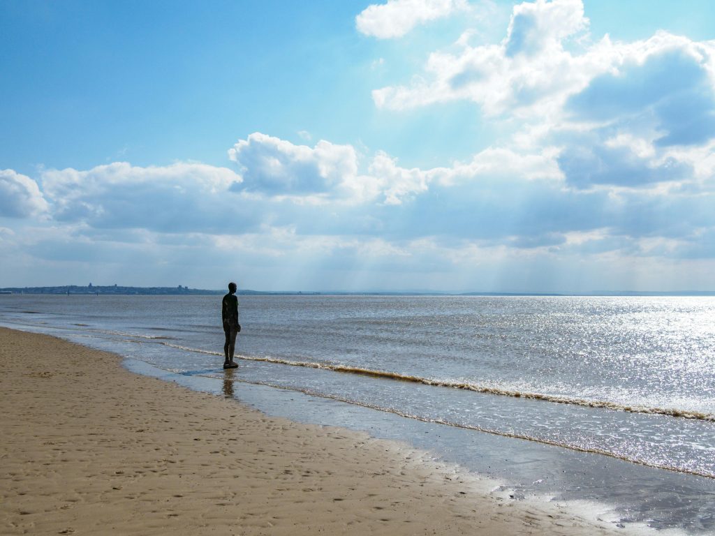 A statue stands alone on Crosby Beach, England under a partly cloudy sky by the sea.